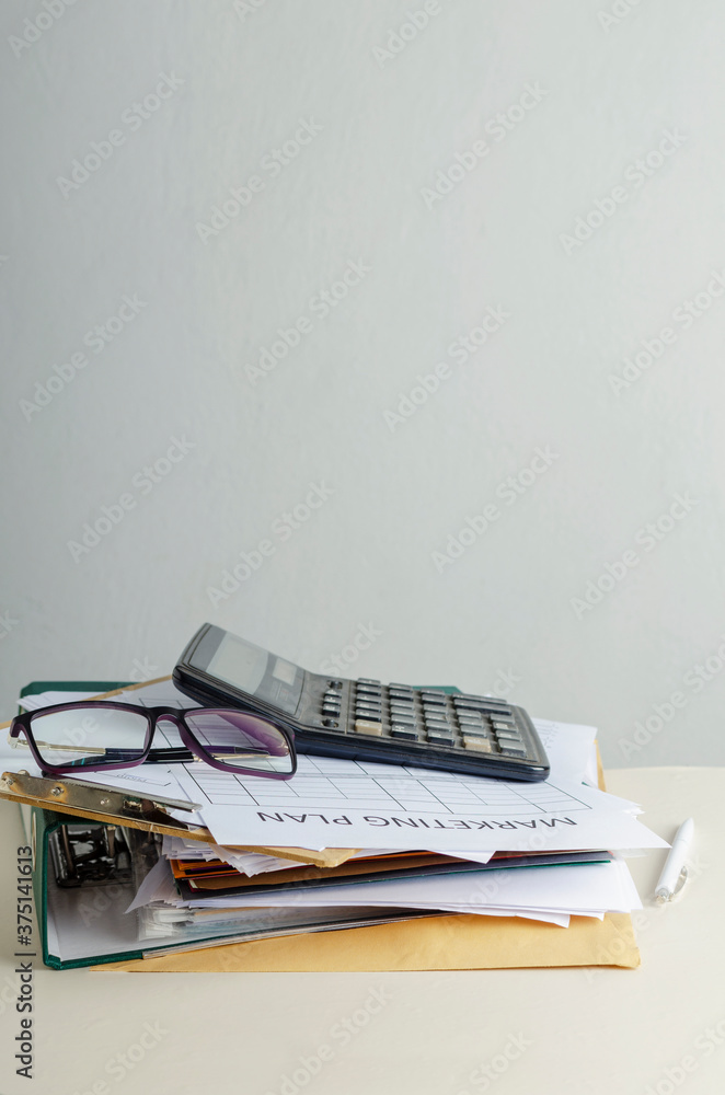 Vertical image. Stack of office documents and blank market plan, glasses, calculator on the work desk against white wall.Empty space