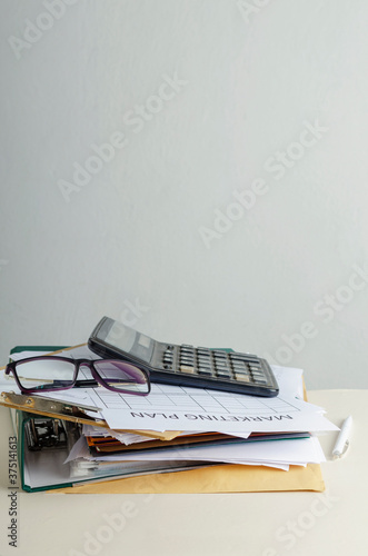 Vertical image. Stack of office documents and blank market plan, glasses, calculator on the work desk against white wall.Empty space