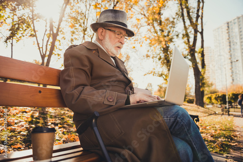 Profile side photo of focused old freelancer work laptop in autumn forest town park chatting searching october weather information sit bench outside wear cap headwear coat