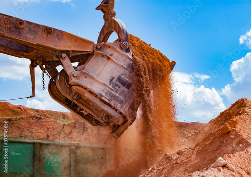 Aluminium ore quarry. Bauxite clay open-cut mining. Loading ore to railway hopper car train with excavator. Close-up of excavator bucket on blue sky with clouds.