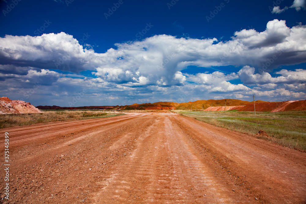 Colorful landscape of bauxite mine (aluminium ore quarry). Orange soil and road and green grass. Blue sky with clouds.
