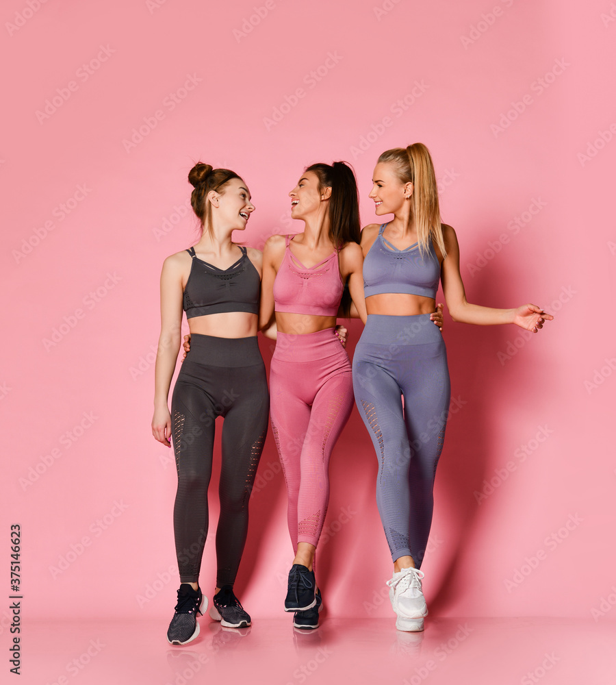 Fototapeta premium Cheerful athletic girls friends in pink, grey, brown training suits walk together chatting. Fitness and yoga girls