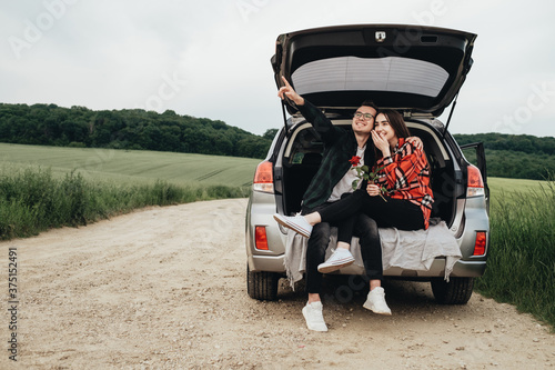 Young Beautiful Couple Sitting in the Car Trunk and Enjoying the Roadtrip, Girl Holding a Rose, Romantic Mood © Romvy