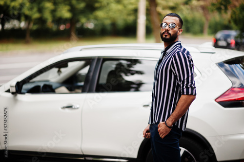 Successful arab man wear in striped shirt and sunglasses pose near his white suv car. Stylish arabian men in transport.