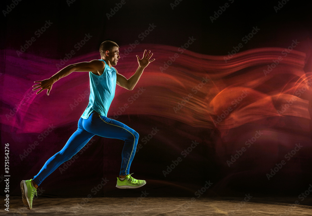 Professional male runner training isolated on black studio background in mixed light. Man in sportsuit practicing in run or jogging. Healthy lifestyle, sport, workout, motion and action concept.