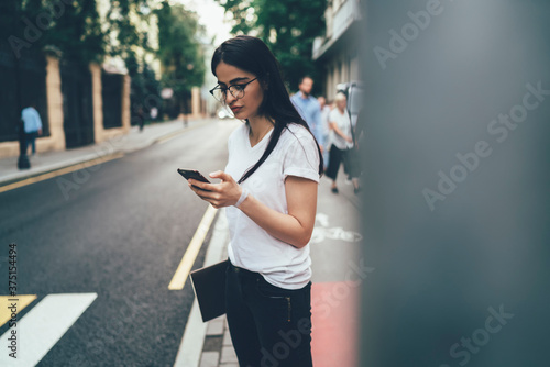 Millennial woman in optical eyewear using internet connection for browsing blog website, Spanish generation Z standing at roadside and checking email message during smartphone online chatting © BullRun