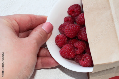 Food delivery. Raspberries in a white plate on a light background. Berries in a woman's hand.