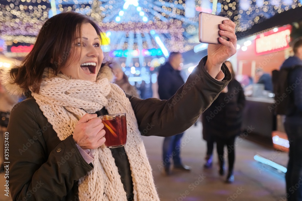 Christmas market, mature happy woman with mug of mulled wine and smartphone taking selfie