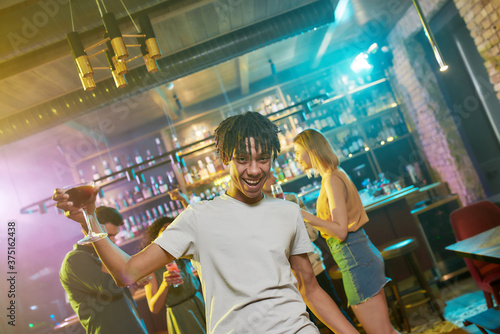  Cheerful mixed race young man getting drunk, smiling at camera with a cocktail in his hand, friends chatting, dancing, having fun in the background