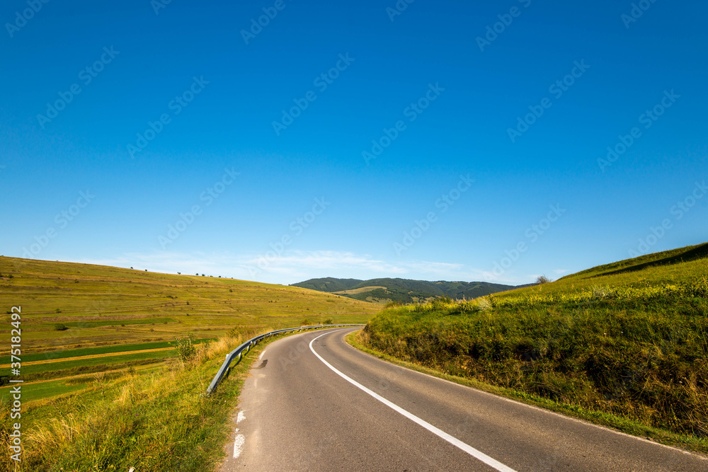 Asphalt road leading through green hills in Transylvania, Romania, blue sky background at early autumn.