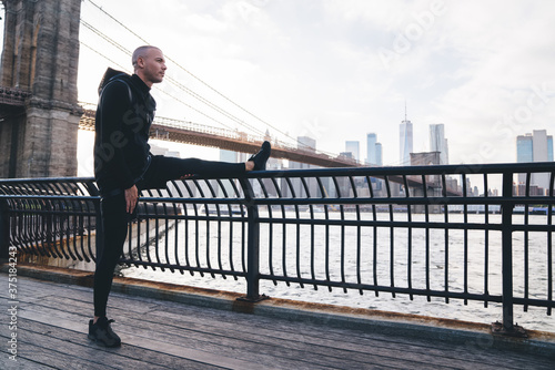 Young ethnic man stretching leg on embankment railing