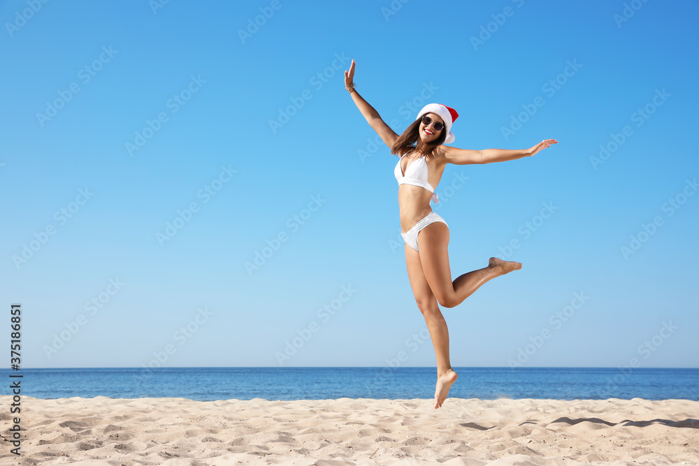 Young woman in Santa hat and bikini jumping on beach, space for text. Christmas vacation