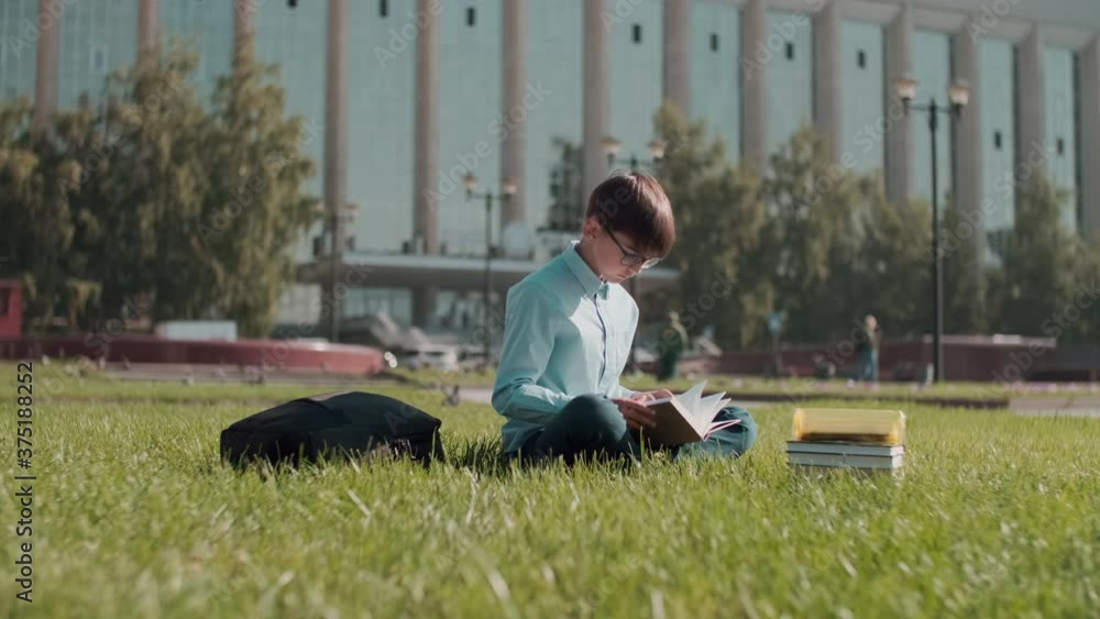 Online education, Back to school, Happy schoolboy, Learn lessons. Schoolboy reading a book while sitting on the grass near the school Online education, Back to school, Happy schoolboy, Learn lessons. Schoolboy reading a book while sitting on the grass near the school