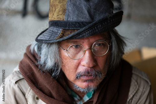 Closeup of old homeless asian man with brown hair, bearded, mustache, glasses, hat and blanket sitting on street in the city.