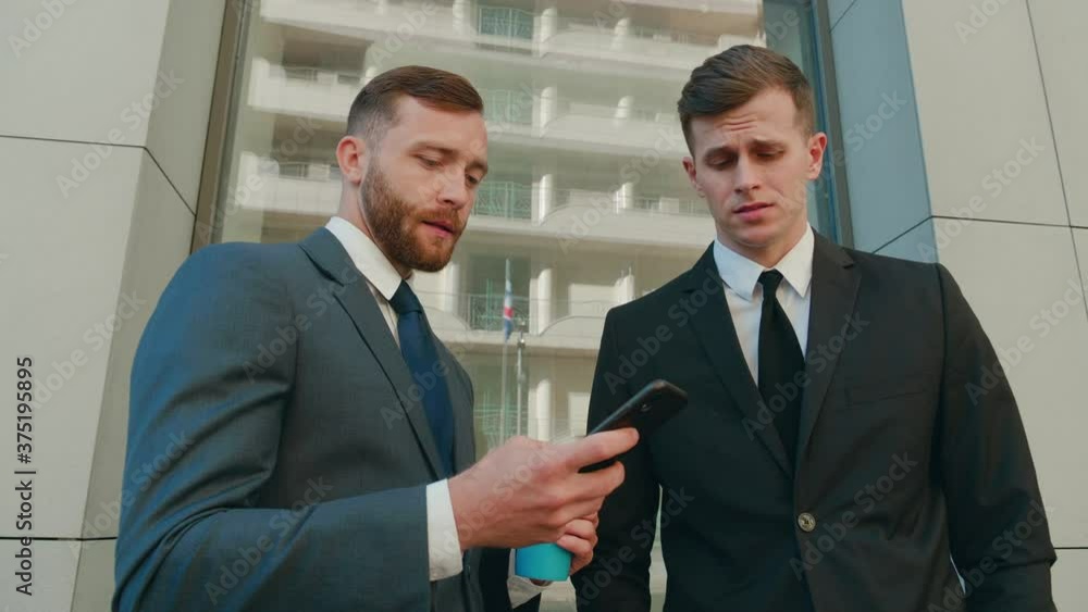 Two male employees interacting with each other in downtown business district. Confident businessmen using smartphone discussing planning computer business project brainstorm in corporate teamwork Two male employees interacting with each other in downtown business district. Confident businessmen using smartphone discussing planning computer business project brainstorm in corporate teamwork