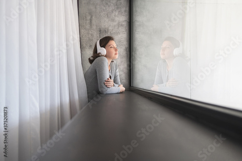 Portrait of beautiful сaucasian young woman in headphones closing eyes dreaming, enjoying listening music standing near big panoramic window of apartments.