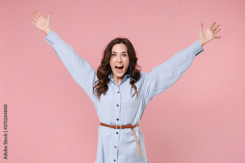Cheerful joyful excited young brunette woman 20s in casual blue shirt dress posing rising spreading hands keeping mouth open looking camera isolated on pastel pink colour background studio portrait. Cheerful joyful excited young brunette woman 20s in casual blue shirt dress posing rising spreading hands keeping mouth open looking camera isolated on pastel pink colour background studio portrait.