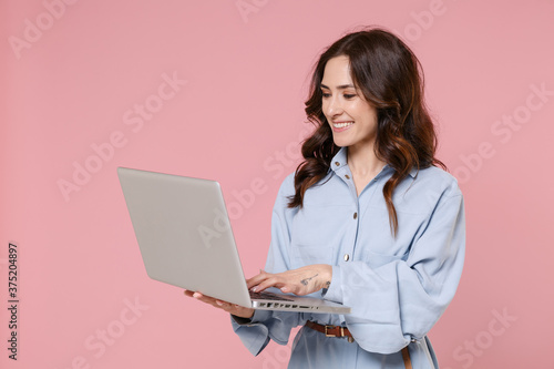 Fotografia Smiling beautiful young brunette woman 20s wearing casual blue shirt dress posing standing hold in hands working in laptop pc computer isolated on pastel pink colour background, studio portrait