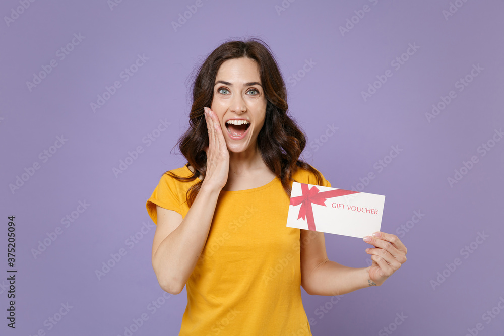 Surprised young brunette woman 20s wearing basic yellow t-shirt posing hold in hands gift certificate put hand on cheek looking camera isolated on pastel violet colour background, studio portrait. Surprised young brunette woman 20s wearing basic yellow t-shirt posing hold in hands gift certificate put hand on cheek looking camera isolated on pastel violet colour background, studio portrait.