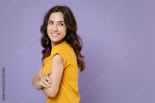 Side view of smiling beautiful young brunette woman 20s wearing basic yellow t-shirt posing standing holding hands crossed looking aside isolated on pastel violet colour background, studio portrait.