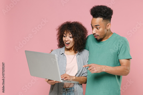 Shocked young african american couple friends guy girl in gray green clothes posing hugging pointing index finger on laptop pc computer isolated on pastel pink color wall background studio portrait.
