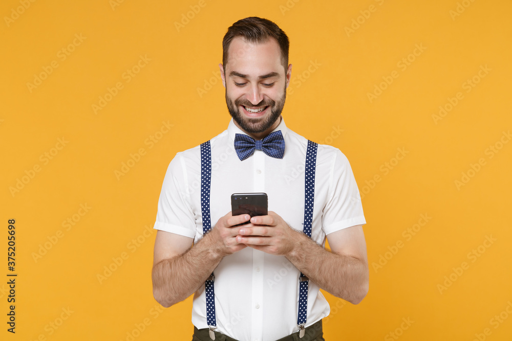 Smiling handsome young bearded man 20s wearing white shirt bow-tie suspender posing standing using mobile cell phone typing sms message isolated on bright yellow color wall background studio portrait. Smiling handsome young bearded man 20s wearing white shirt bow-tie suspender posing standing using mobile cell phone typing sms message isolated on bright yellow color wall background studio portrait.
