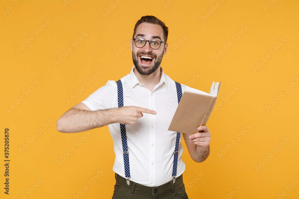 Excited young bearded man 20s wearing white shirt suspender eyeglasses posing standing holding in hands pointing index finger on book isolated on bright yellow color wall background, studio portrait. Excited young bearded man 20s wearing white shirt suspender eyeglasses posing standing holding in hands pointing index finger on book isolated on bright yellow color wall background, studio portrait.