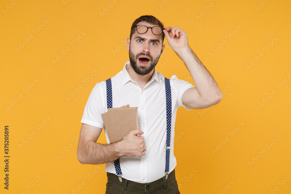 Shocked perplexed worried young bearded man 20s wearing white shirt suspender eyeglasses posing standing hold in hands books looking camera isolated on bright yellow color background, studio portrait. Shocked perplexed worried young bearded man 20s wearing white shirt suspender eyeglasses posing standing hold in hands books looking camera isolated on bright yellow color background, studio portrait.