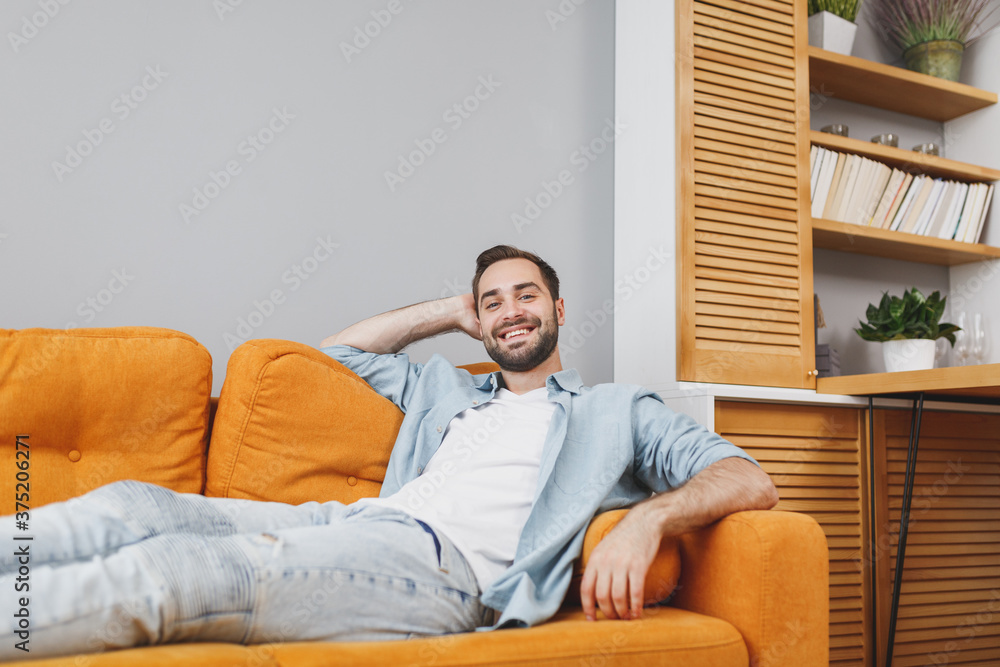 Smiling cheerful handsome young bearded man 20s wearing casual white t-shirt blue shirt looking camera holding hand behind head sitting on couch resting relaxing spending time in living room at home.
