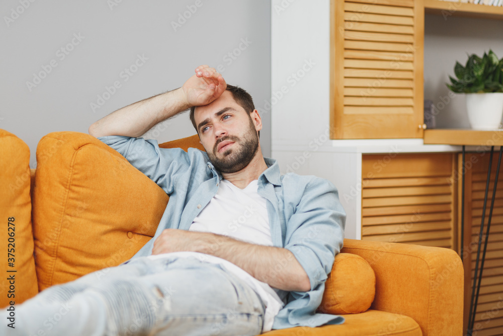 Exhausted tired sick young bearded man wearing casual white t-shirt blue shirt looking aside put hand on head having headache lying on couch resting relaxing spending time in living room at home.