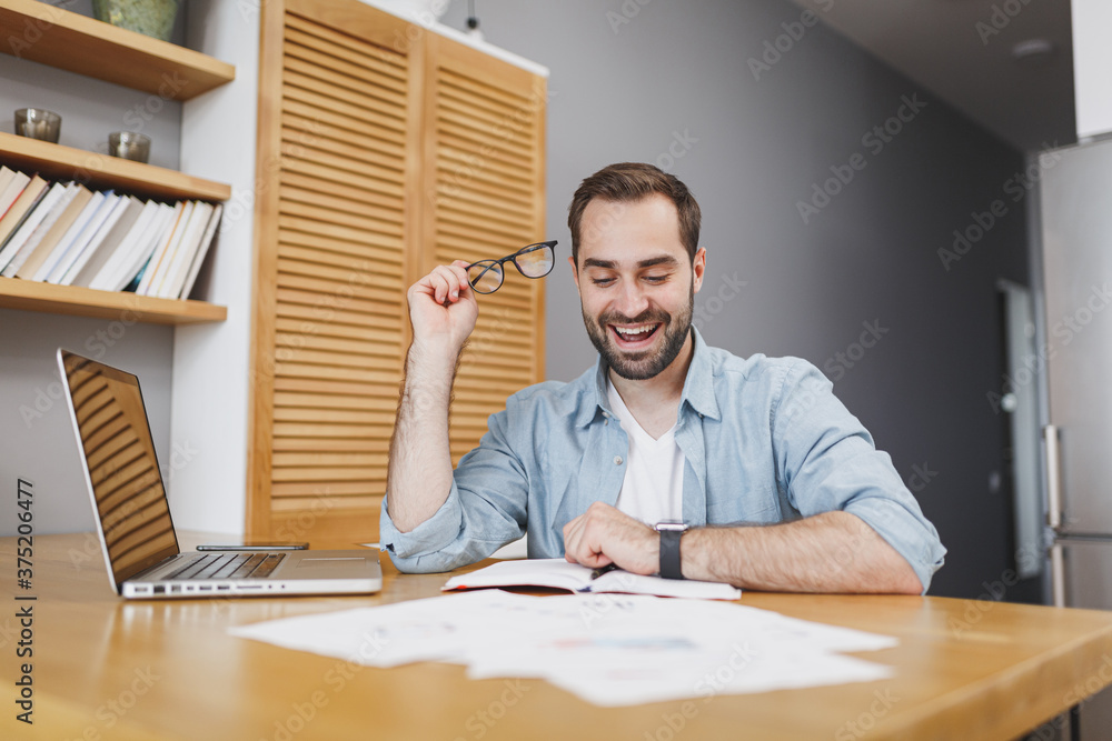 Handsome smiling cheerful funny confident young bearded business man 20s wearing blue shirt glasses sitting at desk with papers document working on laptop pc computer at home or office.