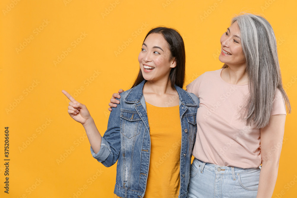 Smiling family asian female women girls mother and daughter in casual clothes posing hugging pointing index finger aside up on mock up copy space isolated on yellow color background studio portrait. Smiling family asian female women girls mother and daughter in casual clothes posing hugging pointing index finger aside up on mock up copy space isolated on yellow color background studio portrait.