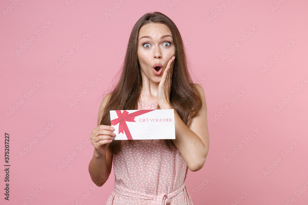 Shocked young brunette woman 20s wearing pink summer dotted dress posing holding in hand gift certificate put hand on cheek looking camera isolated on pastel pink color background studio portrait. Shocked young brunette woman 20s wearing pink summer dotted dress posing holding in hand gift certificate put hand on cheek looking camera isolated on pastel pink color background studio portrait.