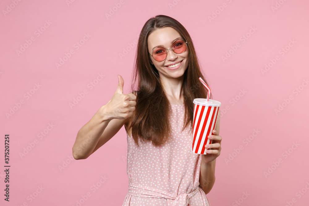 Smiling young brunette woman 20s wearing pink summer dotted dress eyeglasses posing hold plastic cup of cola or soda showing thumb up isolated on pastel pink color wall background studio portrait. Smiling young brunette woman 20s wearing pink summer dotted dress eyeglasses posing hold plastic cup of cola or soda showing thumb up isolated on pastel pink color wall background studio portrait.