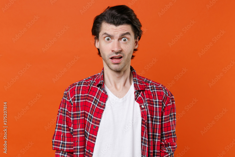 Shocked perplexed worried young brunet man 20s wearing white t-shirt red checkered shirt posing standing keeping mouth open looking camera isolated on orange color wall background studio portrait. Shocked perplexed worried young brunet man 20s wearing white t-shirt red checkered shirt posing standing keeping mouth open looking camera isolated on orange color wall background studio portrait.