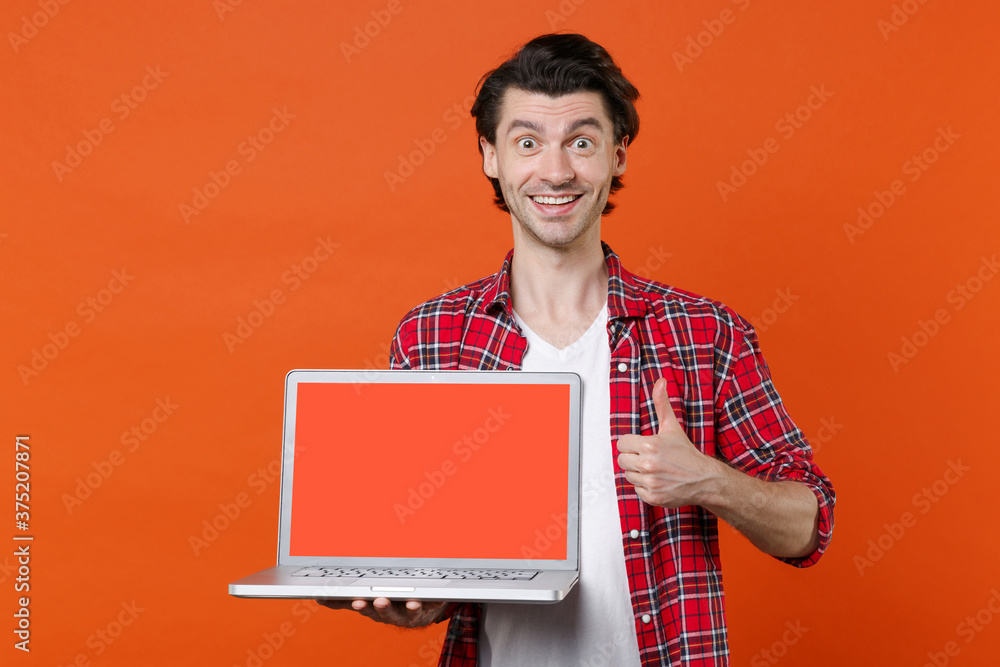 Smiling excited young man 20s in white t-shirt red checkered shirt hold laptop pc computer with blank empty screen mock up copy space showing thumb up isolated on orange background studio portrait.