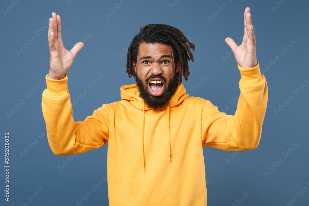 Irritated young african american man guy in yellow streetwear hoodie posing isolated on blue background studio portrait. People sincere emotions lifestyle concept. Spreading hands swearing screaming. Irritated young african american man guy in yellow streetwear hoodie posing isolated on blue background studio portrait. People sincere emotions lifestyle concept. Spreading hands swearing screaming.