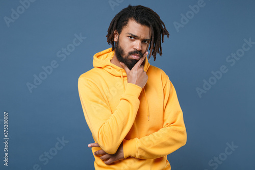 Puzzled pensive young african american man guy wearing yellow streetwear hoodie posing isolated on blue background studio portrait. People sincere emotions lifestyle concept. Put hand prop up on chin. © ViDi Studio