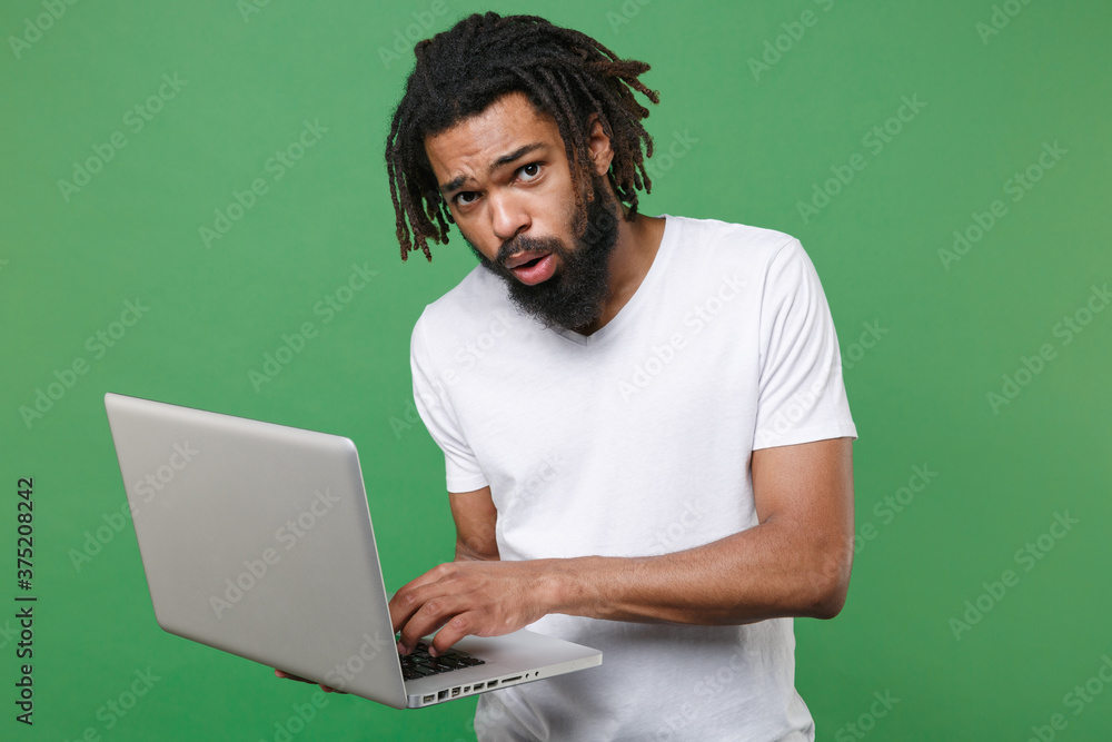 Puzzled shocked worried young african american man guy with dreadlocks 20s wearing white casual t-shirt posing working on laptop pc computer isolated on green color wall background studio portrait.