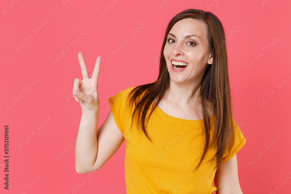 Cheerful funny beautiful smiling young brunette woman 20s wearing yellow casual t-shirt posing standing showing victory sign looking camera isolated on pink color wall background studio portrait. Cheerful funny beautiful smiling young brunette woman 20s wearing yellow casual t-shirt posing standing showing victory sign looking camera isolated on pink color wall background studio portrait.