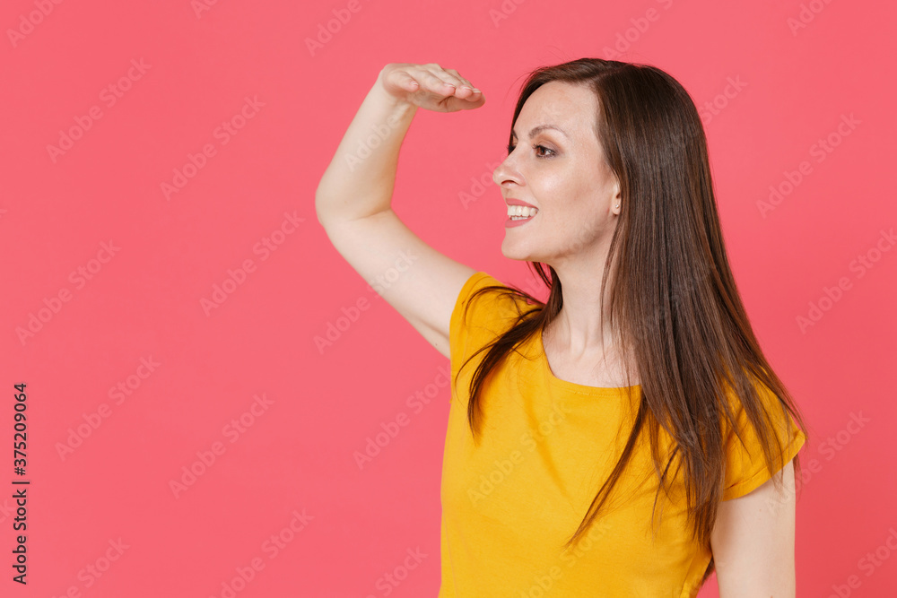 Smiling cheerful funny young brunette woman 20s wearing yellow casual t-shirt posing holding hand at forehead looking far away distance isolated on pink color wall background studio portrait. Smiling cheerful funny young brunette woman 20s wearing yellow casual t-shirt posing holding hand at forehead looking far away distance isolated on pink color wall background studio portrait.