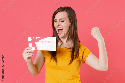 Joyful happy young brunette woman 20s in yellow casual t-shirt posing holding in hand gift certificate doing winner gesture keeping eyes closed isolated on pink color wall background studio portrait.