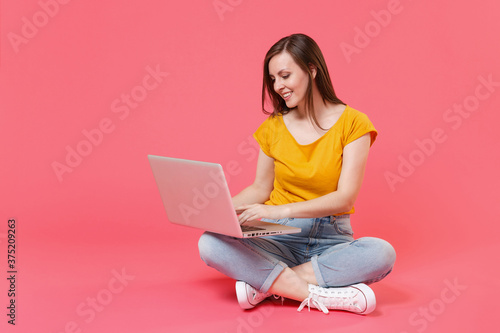 Full length portrait of beautiful smiling attractive young brunette woman 20s wearing yellow casual t-shirt sitting on floor working on laptop pc computer isolated on pink color background studio.