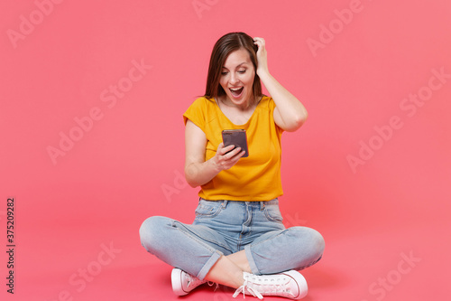 Full length portrait of shocked amazed young brunette woman in yellow t-shirt sit on floor using mobile cell phone typing sms message put hand on head isolated on pink color wall background studio. © ViDi Studio