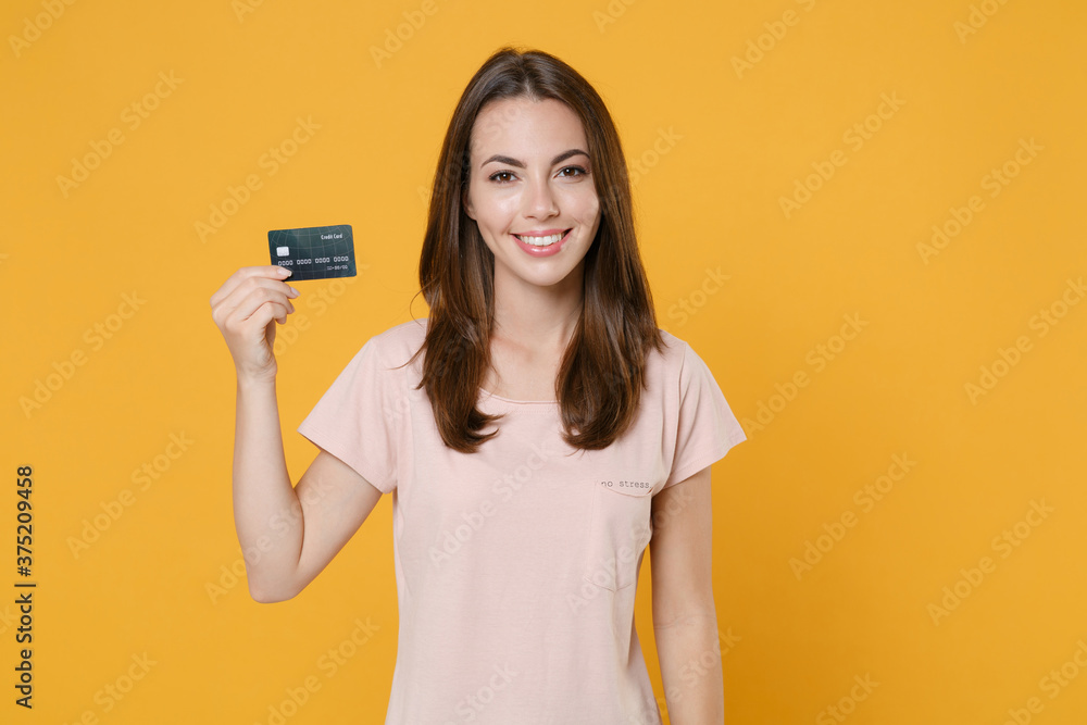 Smiling beautiful young brunette woman 20s wearing pastel pink casual t-shirt posing holding in hand credit bank card looking camera isolated on bright yellow color wall background studio portrait. Smiling beautiful young brunette woman 20s wearing pastel pink casual t-shirt posing holding in hand credit bank card looking camera isolated on bright yellow color wall background studio portrait.