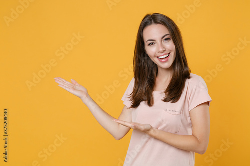 Cheerful laughing young brunette woman 20s in pastel pink casual t-shirt standing posing pointing hands aside on mock up copy space looking camera isolated on yellow color background studio portrait.