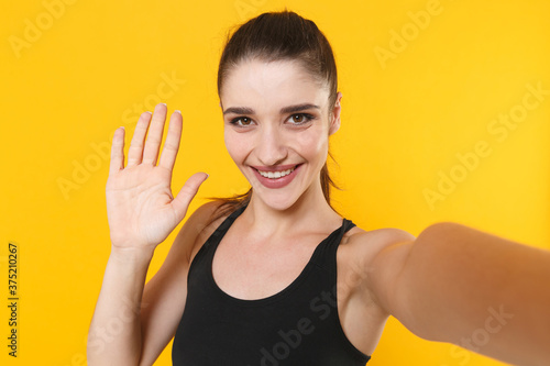 Close up of smiling young fitness sporty woman wearing black sportswear posing training doing selfie shot on mobile phone waving greeting with hand isolated on yellow color background studio portrait. © ViDi Studio