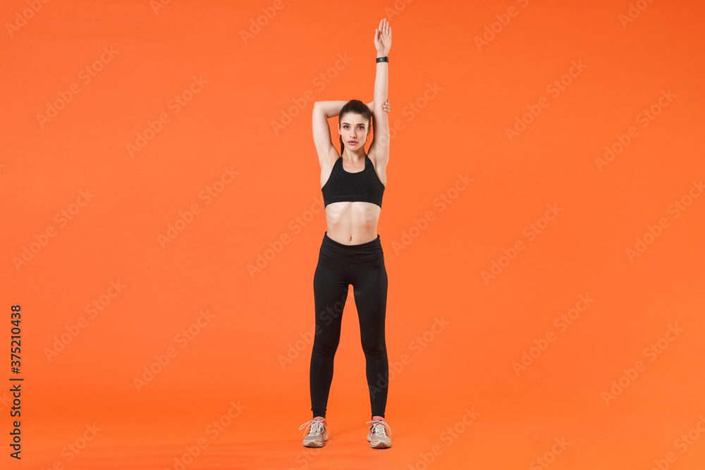 Full length portrait of beautiful young fitness sporty woman in black sportswear posing training working out doing stretching exercising looking camera isolated on orange color wall background studio. Full length portrait of beautiful young fitness sporty woman in black sportswear posing training working out doing stretching exercising looking camera isolated on orange color wall background studio.