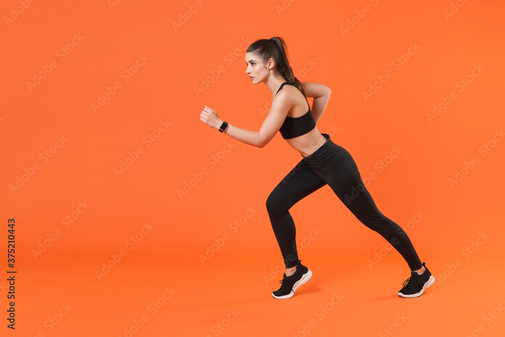 Full length side view portrait of attractive strong young fitness sporty woman 20s wearing black sportswear training working out running looking aside isolated on orange color wall background studio.