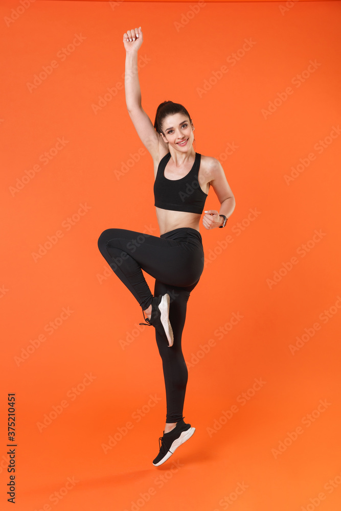 Full length side view portrait of smiling young fitness sporty woman in black sportswear posing training jumping dancing doing cardio exercising rising hands legs isolated on orange background studio.